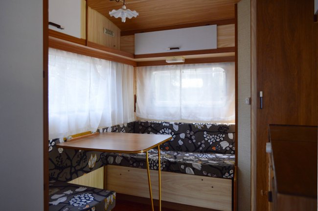 Tavolo e panchette in un camper con finestre luminose e tendine bianche.