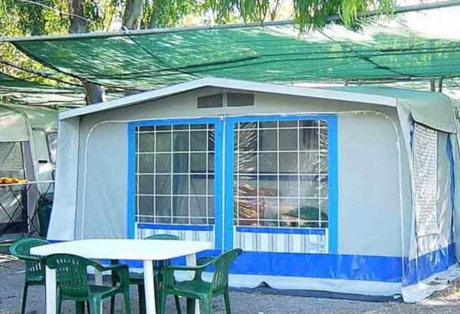Una tenda con struttura rigida, contornata da sedie verdi e un tavolo sotto una rete ombreggiante.
