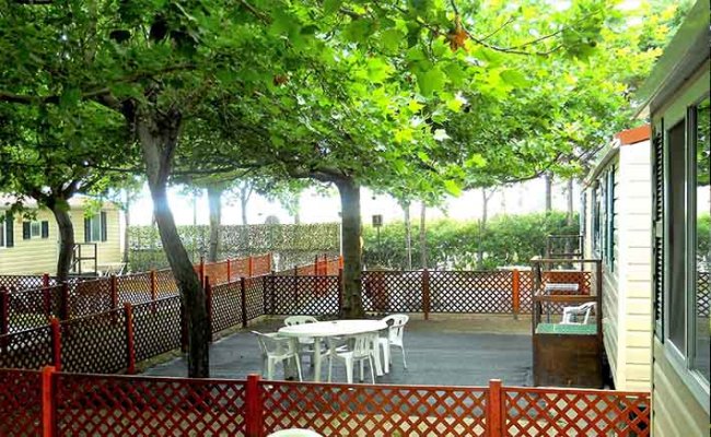 Un cortile con tavoli e sedie bianche, circondato da una staccionata e alberi frondosi.
