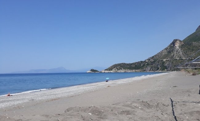 Spiaggia sabbiosa con mare calmo e colline sullo sfondo sotto un cielo azzurro.