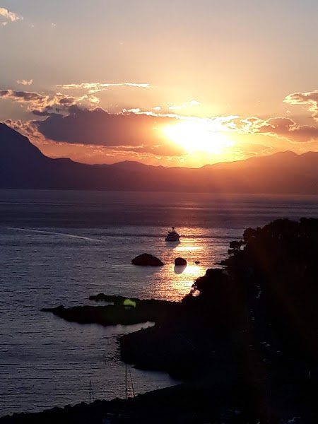 Tramonto sul mare con un'imbarcazione e montagne in lontananza, cielo arancione e nuvole.