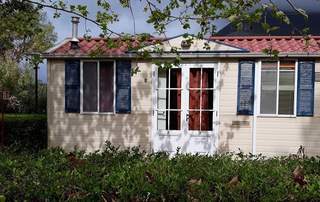 Una piccola casa beige con tetto rosso, finestre con persiane blu e una porta centrale vetrata.