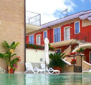 Vista di una piscina con lettini e un edificio rosso a due piani con scale esterne e piante.