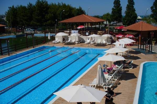 Piscina lunga con corsie, circondata da lettini e ombrelloni bianchi, alberi vicini.