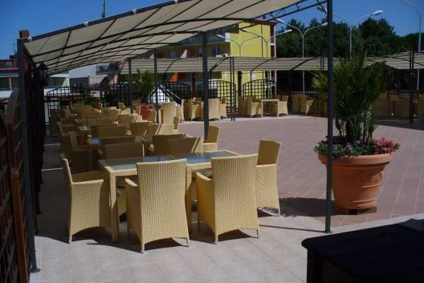 Tavoli con sedie di vimini sotto una tettoia, vasi di piante su una terrazza.