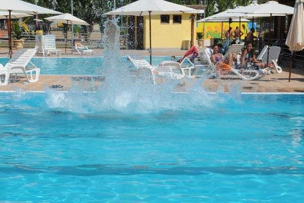 Una piscina con schizzi d'acqua e persone che si rilassano su lettini sotto ombrelloni.