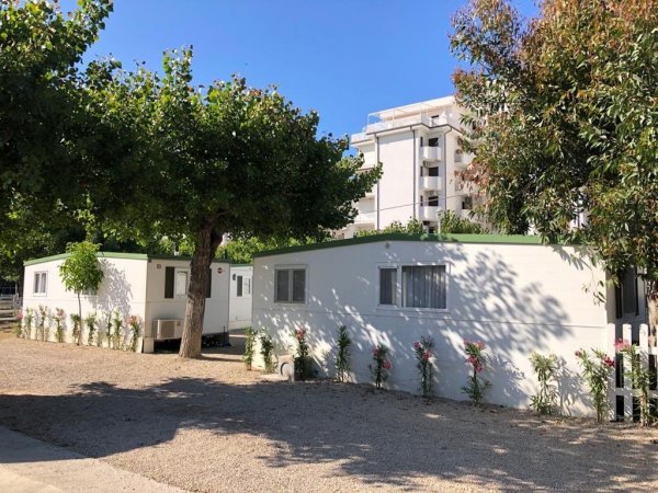 Due case mobili bianche con aiuole, alberi attorno e un edificio sullo sfondo.