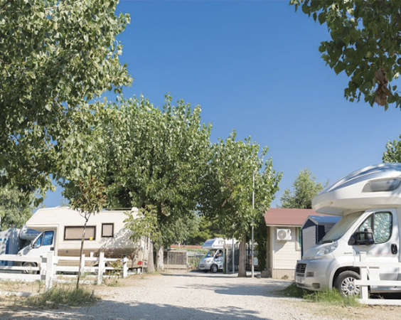 Area camper con veicoli parcheggiati, staccionata bianca e alberi frondosi.