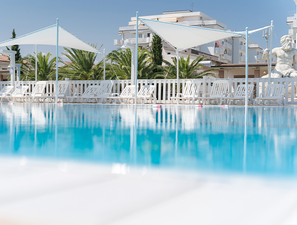 Piscina con sedie a sdraio e vele bianche, edifici sullo sfondo.