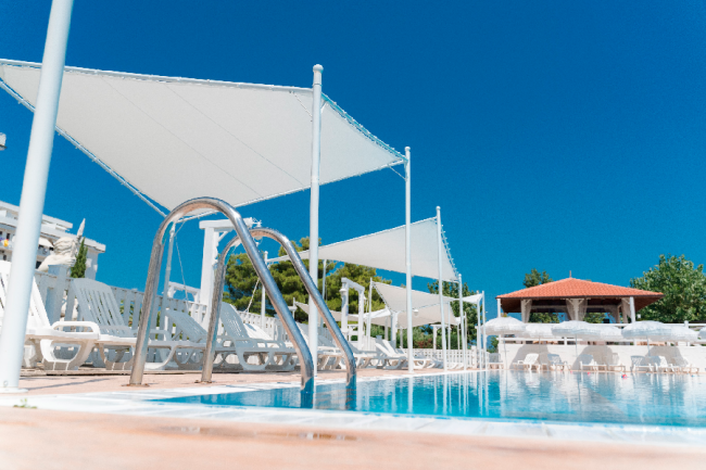 Piscina blu con sedie a sdraio e vele bianche sotto un cielo azzurro.