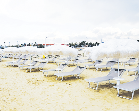 Spiaggia con sabbia chiara, sdraio grigie e ombrelloni bianchi frangiati.