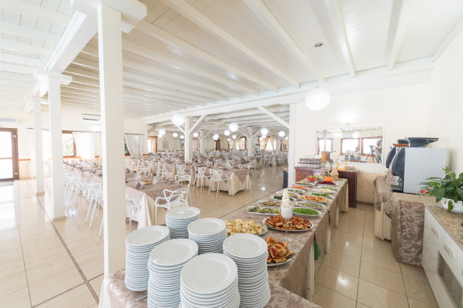 Un buffet con piatti pieni di cibo, vicino a tavoli apparecchiati in una sala da pranzo luminosa.