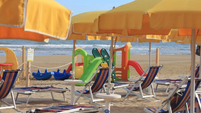 Ombrelloni e lettini gialli su una spiaggia con giochi colorati per bambini in lontananza.