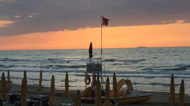 Tramonto sul mare con una bandiera rossa e ombrelloni chiusi in spiaggia.