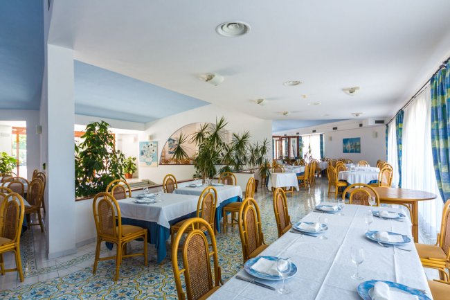 Ristorante con tavoli apparecchiati, sedie in legno e piante decorative.