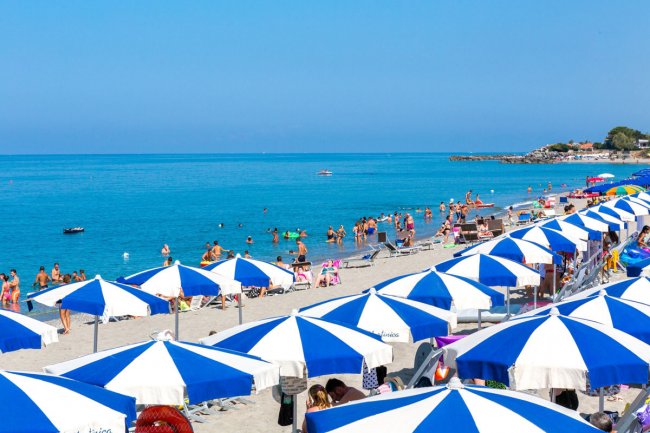 Spiaggia affollata con ombrelloni blu e bianchi, bagnanti in mare e sulla sabbia.