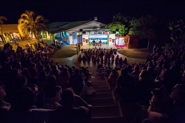 Pubblico seduto all'aperto guarda uno spettacolo serale su un palco.