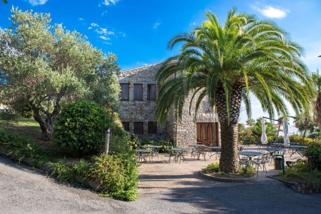 Edificio in pietra con tavolini all'aperto, circondato da palme e vegetazione.