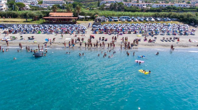 Spiaggia affollata con ombrelloni blu, persone che nuotano e un mare limpido.
