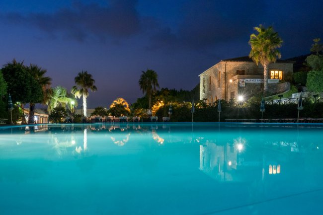 Piscina illuminata di sera con palme e edificio in pietra sullo sfondo.
