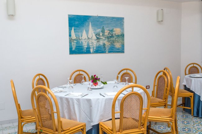 Tavolo rotondo apparecchiato con sedie intrecciate, quadro nautico sul muro.