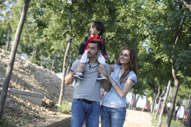 Una famiglia passeggia, con un bambino seduto sulle spalle di un adulto, in un parco alberato.