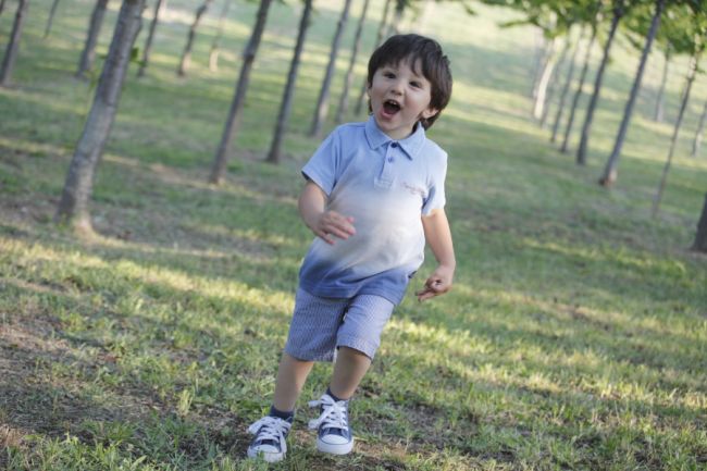 Bambino sorridente corre su un prato con alberi e luce del sole.