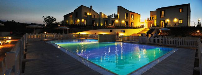 Piscina illuminata di sera, circondata da edifici residenziali e luci calde.