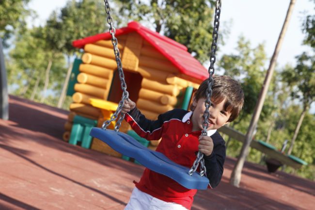 Un bambino su un'altalena blu in un parco giochi con una casetta di legno sullo sfondo.