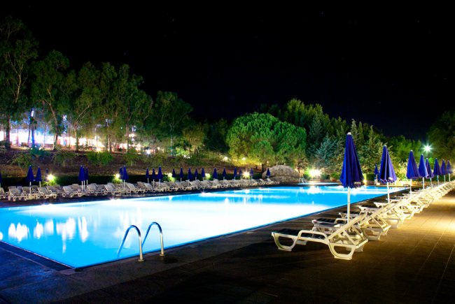 Piscina illuminata di notte, circondata da sdraio bianche e ombrelloni blu.