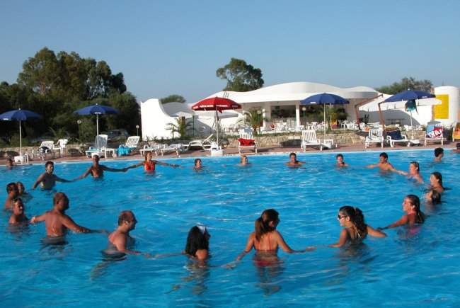 Persone in cerchio in una piscina all'aperto con ombrelloni e un edificio bianco dietro.