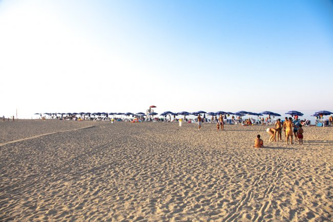 Spiaggia sabbiosa con ombrelloni blu e bagnanti sotto un cielo limpido.
