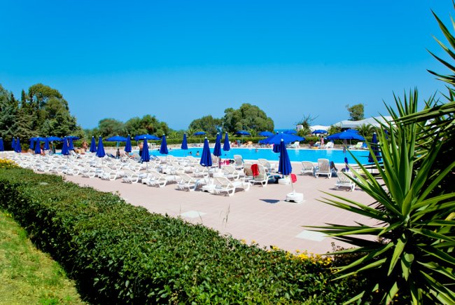Piscina con sedie a sdraio bianche, ombrelloni blu e vegetazione verde intorno.