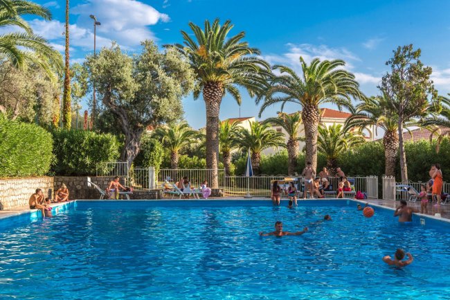Persone che nuotano in una piscina circondata da palme sotto un cielo blu.