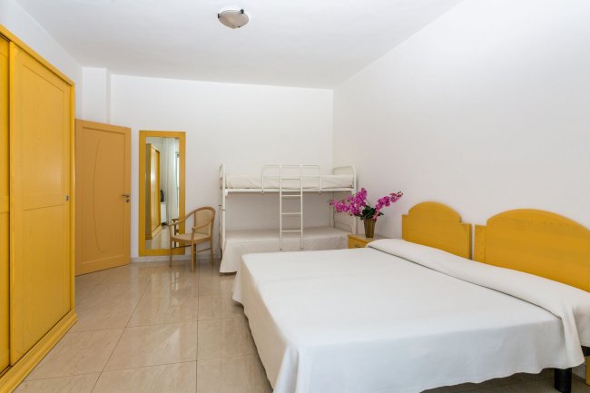 Camera con letto matrimoniale, letto a castello, armadio e sedia. Pareti bianche e mobili gialli.