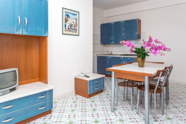 Cucina open space con mobili blu, tavolo da pranzo con fiori rosa e pavimento piastrellato.