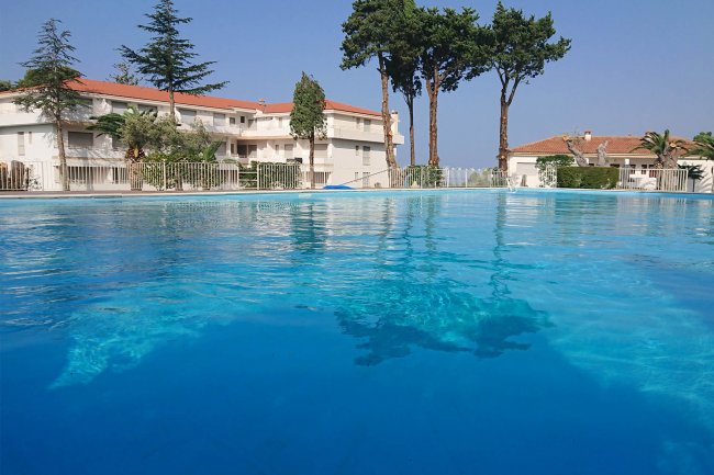 Piscina all'aperto con villette bianche e tetti rossi sullo sfondo, alcune palme e pini.
