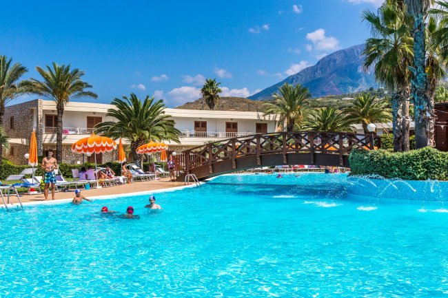Piscina con persone, palme e una passerella in legno, montagne sullo sfondo.