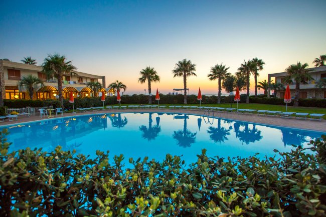 Piscina riflettente al tramonto con palme e strutture sullo sfondo.