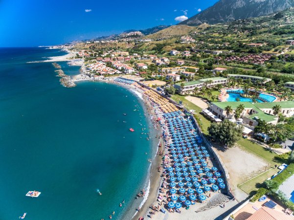 Vista aerea di una spiaggia affollata, ombrelloni allineati e montagne sullo sfondo.
