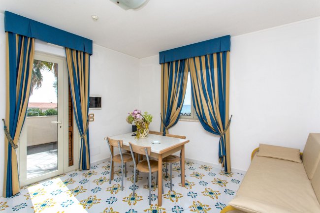 Stanza luminosa con tavolo, sedie, divano, tende blu e porta-finestra verso il balcone.