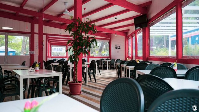 Sala da pranzo interna con tavoli e sedie nere, pareti rosse e grandi finestre.