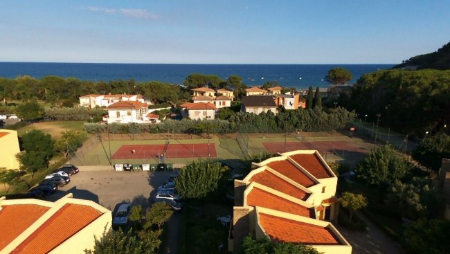 Vista dall'alto di case con tetti rossi, campi da tennis e mare.