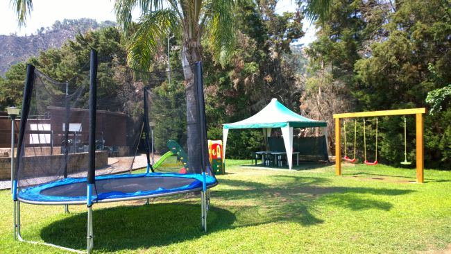 Area giochi con tappeto elastico, altalene e un gazebo su un prato verde.