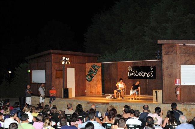 Spettacolo di cabaret serale su un palco di legno con pubblico seduto all'aperto.