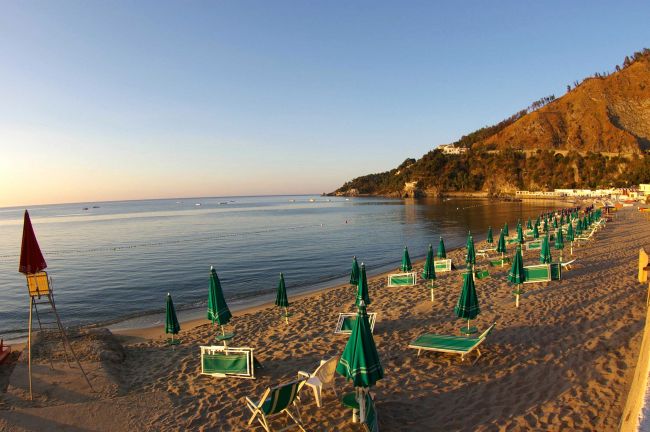 Un tramonto illumina una spiaggia sabbiosa con file di ombrelloni verdi chiusi e sdraio.