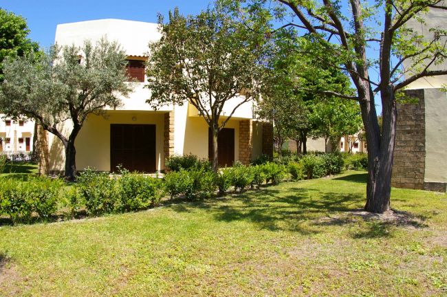 Edificio beige a due piani con grandi porte di legno, circondato da alberi e cespugli verdi.