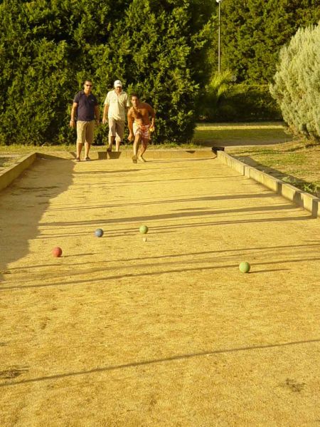 Tre persone giocano a bocce su un campo di sabbia, circondato da alberi.