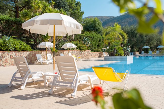 Lettini e ombrelloni bianchi accanto a una piscina azzurra, con vegetazione intorno.