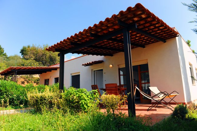 Bungalow con tetto a tegole, sedie in legno e sdraio su un patio circondato da vegetazione.